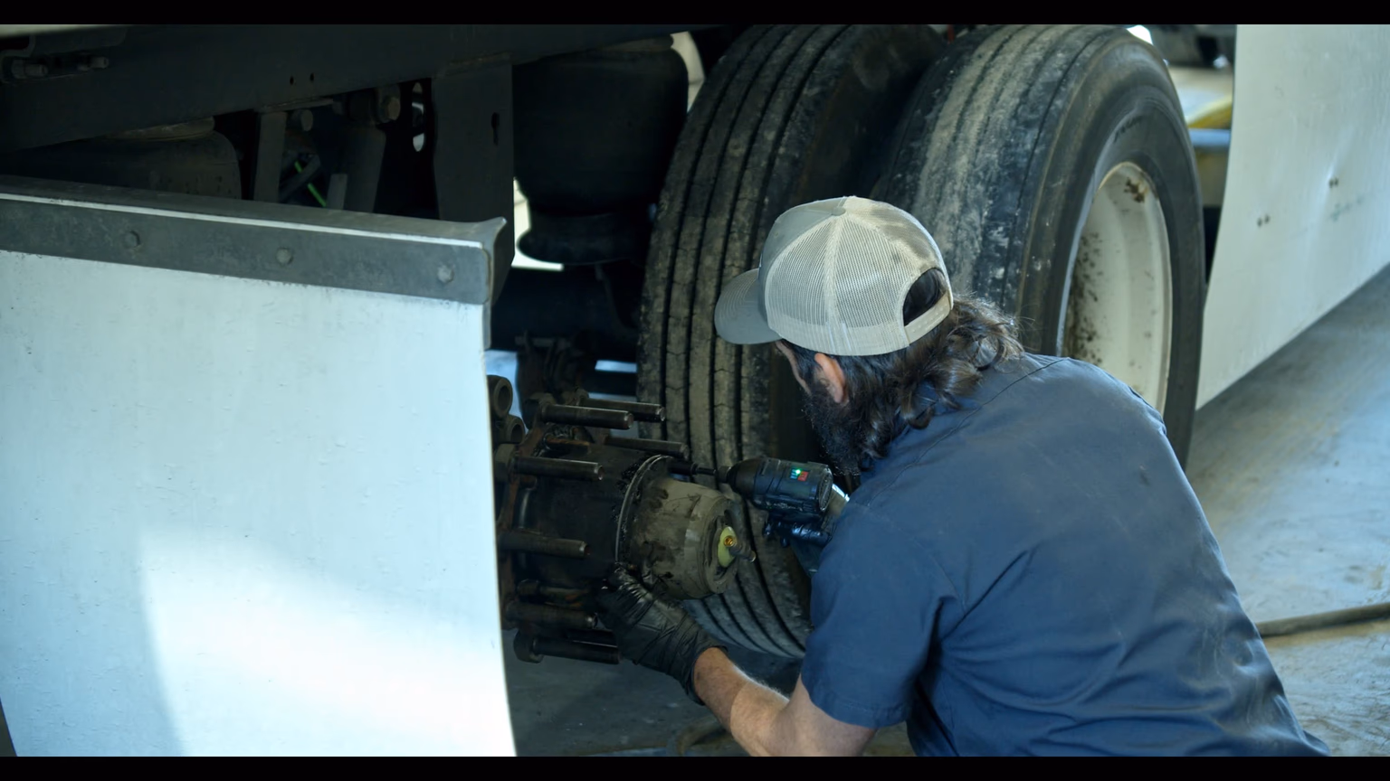 An Onyx Logistics mechanic in coveralls servicing a wheel hub on a Freightliner tractor — hub fully exposed with lug studs visible, powered impact wrench in hand mid-action.