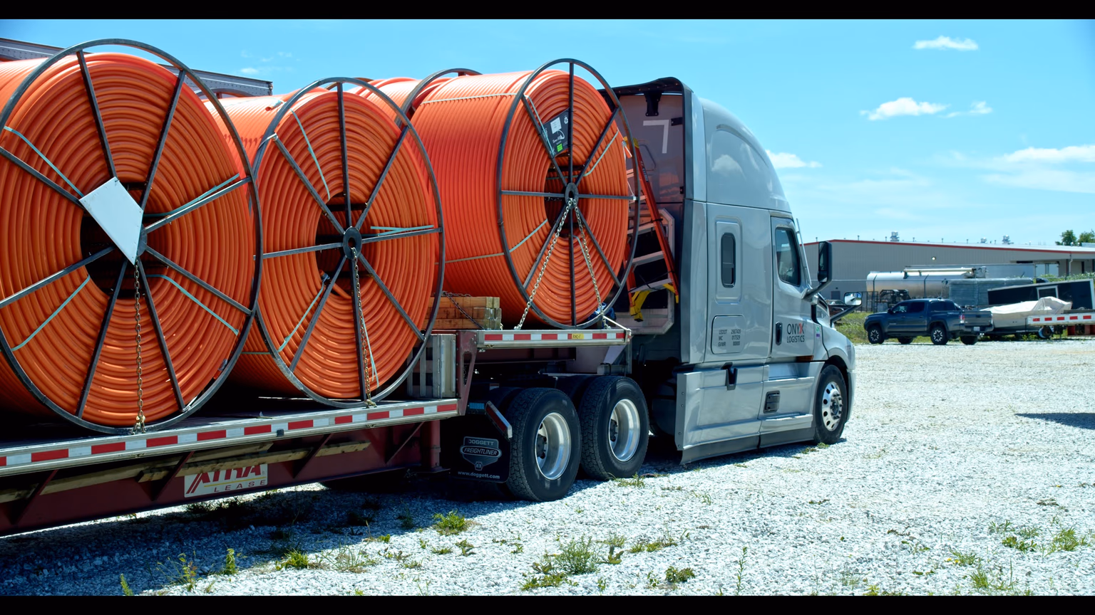 An Onyx Logistics flatbed trailer loaded with three large orange HDPE conduit reels, side-on, under a clear sky.