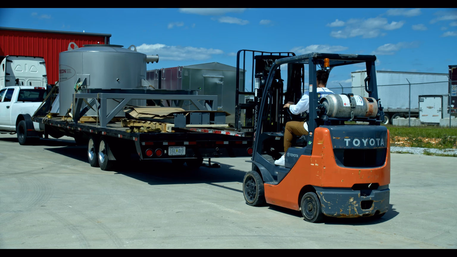 A forklift operator unloading freight from an Onyx Logistics hotshot gooseneck trailer at a delivery destination — Toyota lift truck mid-action approaching the loaded silver tank.